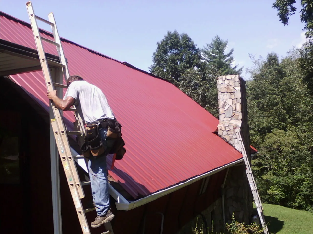 Roofer on a ladder beside a red metal roof and stone chimney — Soffit & Fascia Repair by Roofing Brothers in Maquoketa